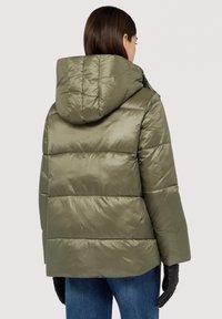 Grön vadderad jacka med huva, med ett quiltat mönster, blank yta och sidofickor. Bärs över blå jeans och svarta handskar.