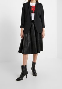 Žena v černém saku, bílém tričku s potiskem, černé kožené midi sukni a černých lodičkách s kotníkovým páskem stojí na bílé podlaze.