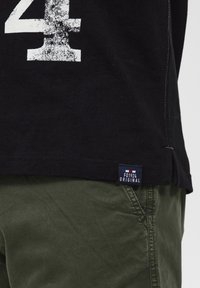 Melnas kokvilnas T-krekls ar nolietotu baltu "4" druku un birku ar uzrakstu "FQ1924 ORIGINAL," apvienots ar olīvu zaļām biksēm.