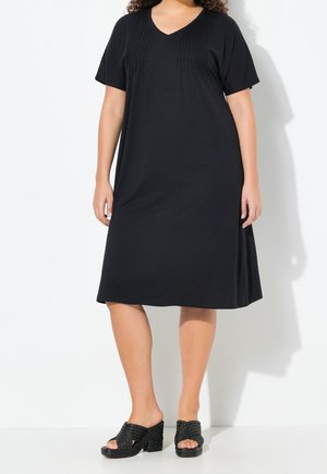 Femme portant une robe noire mi-longue à manches courtes avec des détails plissés près de l'encolure, associée à des sandales noires à talons ouverts.