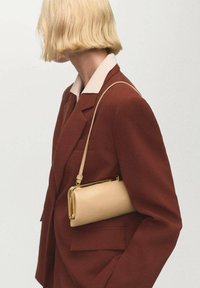 Mujer con cabello corto rubio, vistiendo una chaqueta marrón y llevando un pequeño bolso de hombro beige con un broche dorado.