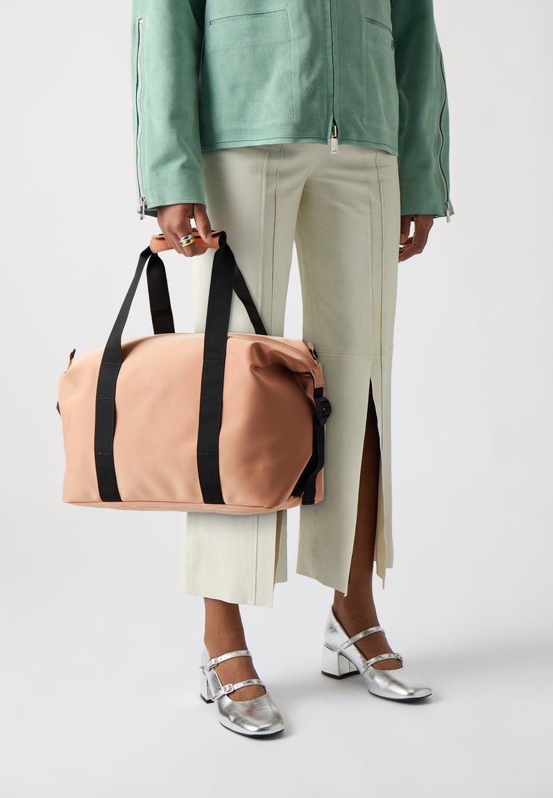 Peachfarget duffelbag med svarte stropper, laget av glatt materiale. Har glidelås og en rund design for enkel håndtering.