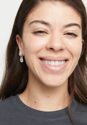 Boucles d'oreilles argentées en forme de goutte avec un design lisse et arrondi. Gros plan du visage mettant en valeur les boucles d'oreilles contre une chemise gris foncé et une texture de peau naturelle.