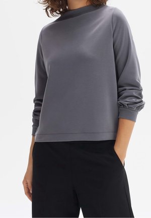 Femme portant un haut gris ample à manches longues froncées et un pantalon noir, une main dans la poche, sur un fond neutre.