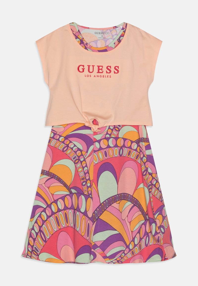Brzoskwiniowy top z krótkim rękawem z czerwonym napisem "GUESS LOS ANGELES", związany w talii, nałożony na kolorową, wzorzystą sukienkę bez rękawów w odcieniach fioletu, pomarańczu, różu i zieleni.