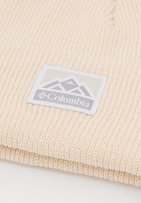 Svetlo béžová pletená čiapka s rebrovým vzorom. Obsahuje obdĺžnikový sivý nášivku s horami a logom "Columbia".