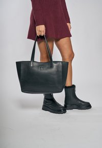 Borsa tote in pelle nera con doppi manici, forma rettangolare, logo impresso, texture liscia, abbinata a stivali neri con zip.