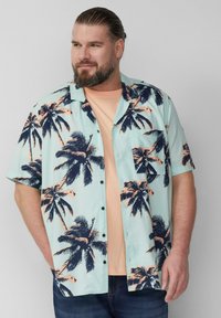 Korte mouwen shirt in lichtblauw met donkere palmboomprint; beschikt over een knoopsluiting aan de voorkant en een borstzak. Gelaagd over een licht perzikkleurig t-shirt.