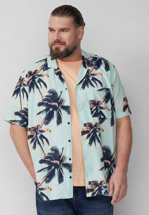 Korte mouwen shirt in lichtblauw met donkere palmboomprint; beschikt over een knoopsluiting aan de voorkant en een borstzak. Gelaagd over een licht perzikkleurig t-shirt.