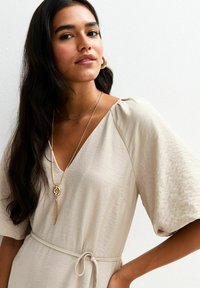 Beige, lichtgewicht jurk met een V-hals, pofmouwen en een taille met strik. Geaccentueerd met gelaagde gouden kettingen en ronde oorbellen.