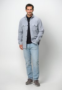 Grå skjorta med knappar och två bröstfickor, svart polo under, ljusblå denimjeans och grå sneakers med vita sulor.