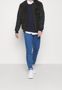 Man som bär en kamouflagemönstrad fleecejacka, vit skjorta, marinblå tröja, blå upprullade jeans och vita sneakers, går mot en enfärgad bakgrund.