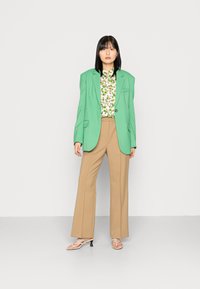 Blazer verde ajustado sobre uma blusa branca com padrões florais, combinado com calças largas bege e sandálias strappy bege. Texturas de tecido suaves.