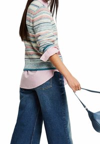 Femme portant un pull en tricot rayé sur une chemise rose et un jean large, tenant un sac à main bleu par sa sangle contre un fond blanc.