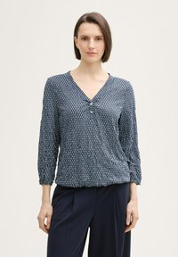 Blouse marine avec un motif géométrique, décolleté en V et manches trois-quarts. Présente une fermeture à boutons et des détails froncés au niveau des manches.