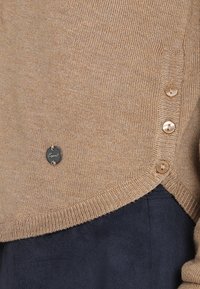 Pull en maille marron avec une fermeture à boutons sur le côté, comprenant cinq boutons ronds et une petite étiquette logo en métal à l'ourlet.
