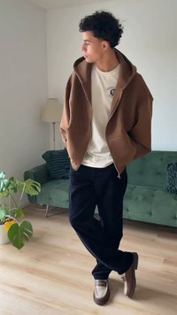 Brązowa bluza z zamkiem na suwak na białej koszulce, luźne czarne spodnie i brązowe mokasyny z kremowymi akcentami, stojący w jasnym pokoju z zieloną kanapą.