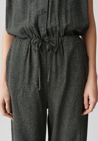 Grüner Jumpsuit mit gemustertem Design, das eine strukturierte, abstrakte Optik aufweist. Er verfügt über einen Kordelzug in der Taille und dunkle Knöpfe zum Verschließen.