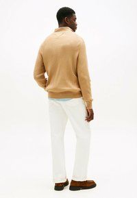 Uomo di spalle che indossa un maglione beige a coste, pantaloni bianchi, camicia azzurro chiaro sotto e scarpe di pelle scamosciata marroni, su sfondo bianco.