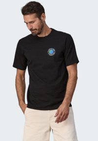 Zwarte katoenen T-shirt met korte mouwen, voorzien van een kleurrijk rond logo op de linkerborst, gedragen met lichtgekleurde shorts.