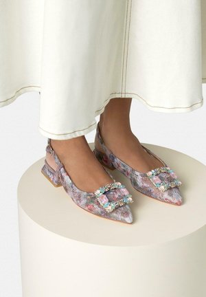 Füße in floralen Spitzen-Slingback-Ballerinas mit quadratischen, mit Edelsteinen besetzten Schnallen, kombiniert mit einem cremefarbenen langen Rocksaum.