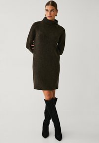 Dunkelbraunes, geripptes Strickkleid mit hohem Rollkragen und langen Ärmeln, gerader Schnitt und knielange Gestaltung. Es werden schwarze kniehohe Stiefel getragen.