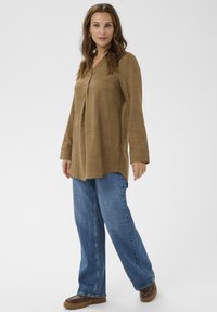 Túnica de manga larga en marrón claro con cuello y detalle de pliegue, combinada con jeans azules de pierna ancha y zapatos marrones. Tela texturizada.