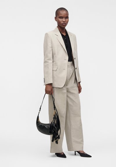 Personne portant un costume beige avec un pantalon à jambes larges et un blazer, une chemise noire, des talons noirs, tenant un sac à main noir avec des détails en pompons.