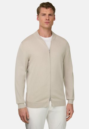 Uomo con cardigan beige con zip sopra una camicia bianca e pantaloni bianchi, in piedi davanti a uno sfondo chiaro uniforme.