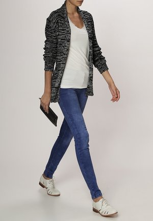 Un cardigan en tricot noir et blanc porté par-dessus un haut blanc, associé à un jean skinny bleu et des chaussures blanches. La personne tient une pochette noire.
