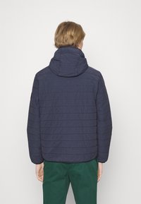 Giacca imbottita blu navy con righe di trapuntatura orizzontale, colletto alto e cappuccio regolabile, abbinata a pantaloni verdi. Texture liscia.
