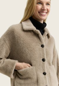 Beige fuzzy jas met een ingesneden kraag, zwarte knopen en zijzakken. De textuur van de stof lijkt zacht en warm te zijn.