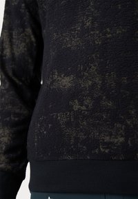 Fleece puloveris tumši melnā krāsā ar faktūru. Iezīmēts ar ribotām piedurknēm un apakšmalu, kā arī kontrastējošām gaišām svītrām visā audumā.