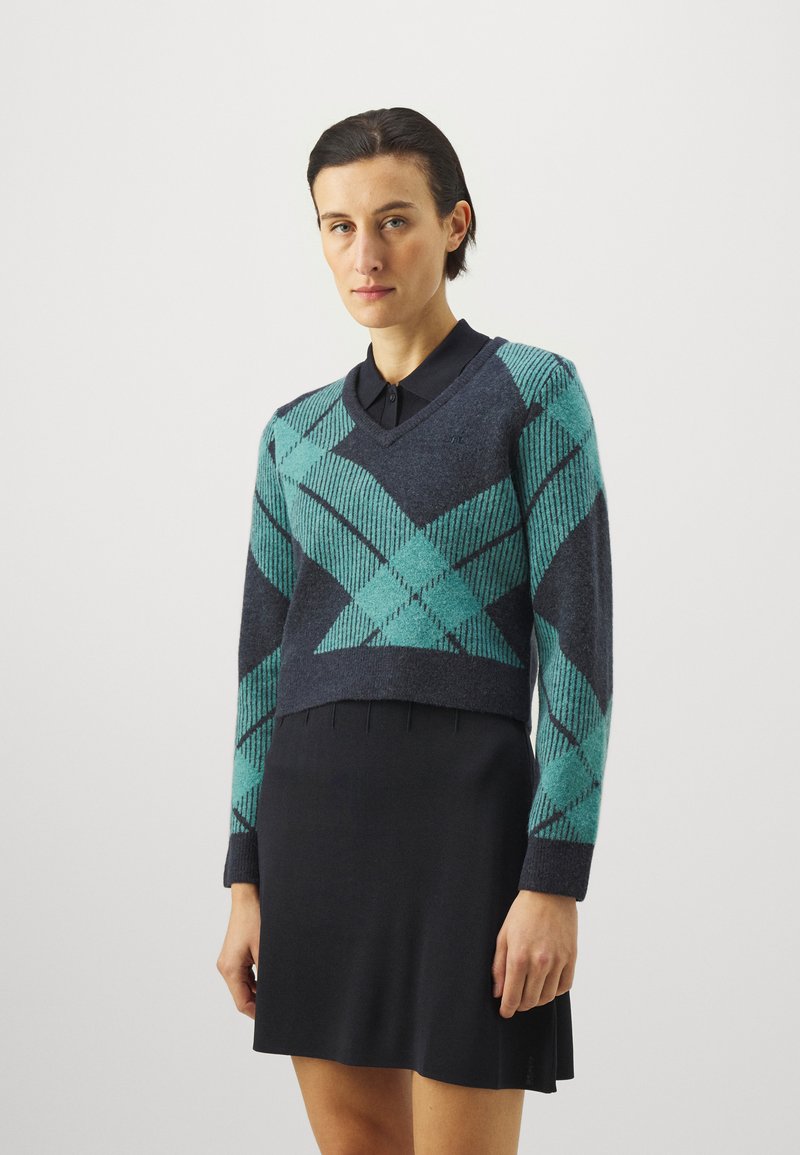 Avkuttet genser i marineblå med teal argyle-mønster, med V-hals og lange ermer, over en svart kjole med frynser nederst.