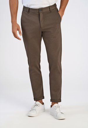 Braune maßgeschneiderte Hosen aus glattem Stoff, mit Knopfverschluss, Seitentaschen und aufgerollten Säumen, kombiniert mit weißen Sneakers.