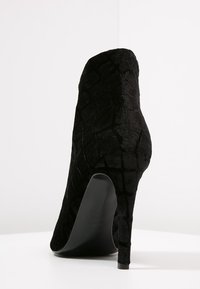 Botín negro de terciopelo con un patrón texturizado, puntera afilada y un alto tacón de aguja, que muestra una silueta suave y un diseño elegante.