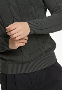 Maglione a maglia grigio scuro con design a trecce, polsini a coste e orlo a coste. Le mani stanno regolando il polsino.
