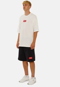 Weißes T-Shirt mit kurzen Ärmeln und rotem Logo, kombiniert mit schwarzen Shorts mit einem roten Patch, abgerundet mit weißen Turnschuhen.