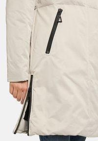 Manteau pour femme gris clair avec une finition lisse, doté d'accents de fermeture éclair noirs, d'une poche latérale et d'un ourlet évasé pour faciliter le mouvement.