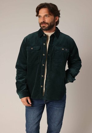 Donkergroene corduroy shirtjack met twee voorkruizen, knoopsluiting en een zachte textuur, gedragen over een crèmekleurige trui en blauwe spijkerbroek.