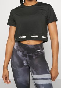 Camiseta corta negra con texto blanco "STRONGER" en el dobladillo, combinada con leggings de cintura alta que tienen un patrón de tie-dye en negro y gris y acentos a rayas.
