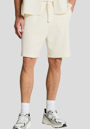 Man draagt crèmekleurige gebreide shorts, witte sokken tot aan de kuit en witte en zwarte sportschoenen, staand met één hand in de zak.