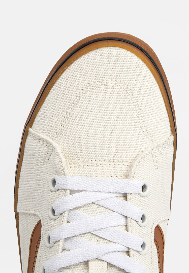 Zapatilla de lona en color blanco sucio con acentos marrones, cordones blancos y suela de goma. Presenta una textura suave y un diseño clásico.