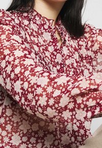 Blusa trasparente rossa con fiori, con placca arricciata, dettaglio dei bottoni e collo alto. Presenta motivi floreali bianchi e rosa chiaro. Maniche lunghe.