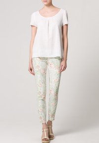 Top blanco de manga corta con detalle de puntilla, combinado con pantalones de estampado floral en verde claro. El modelo lleva sandalias de tiras beige.