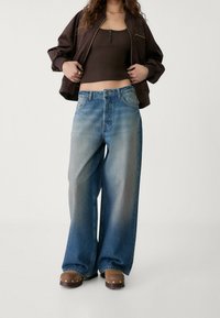 Szerokie, niebieskie dżinsy z wyblakłym gradientem, posiadające tradycyjny pięć kieszeni, wysoki stan i zapięcie na guzik. Pokazane brązowe drewniane klapki.