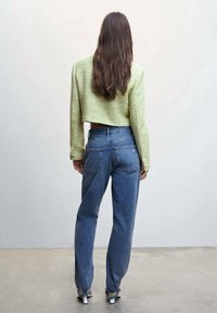 Donna con lunghi capelli castani indossa un giacchino verde a coste, jeans blu a vita alta e tacchi bianchi, in piedi davanti a uno sfondo neutro.