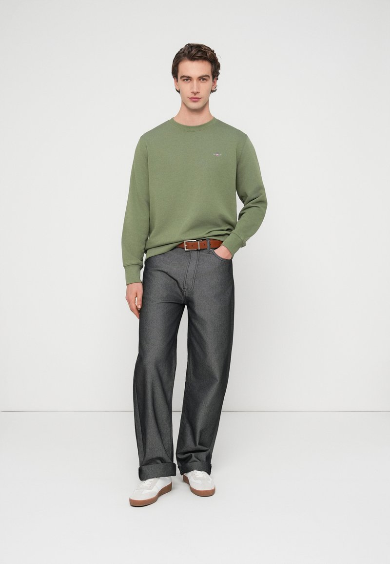 Pull en coton vert, jean gris à jambes larges, ceinture en cuir marron et baskets blanches. Tenue décontractée avec manches ajustées et revers roulés.