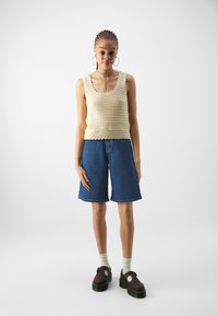 Crop top en laine crème avec des bords festonnés, associé à un short en denim bleu. Le modèle porte des chaussures marron et des chaussettes claires.