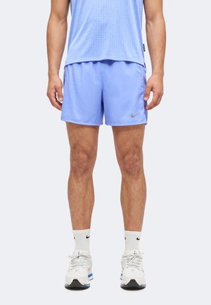 Homme portant un short de sport bleu clair et un débardeur à perforations assorti, chaussettes blanches Nike et chaussures de course blanches, debout, bras détendus.